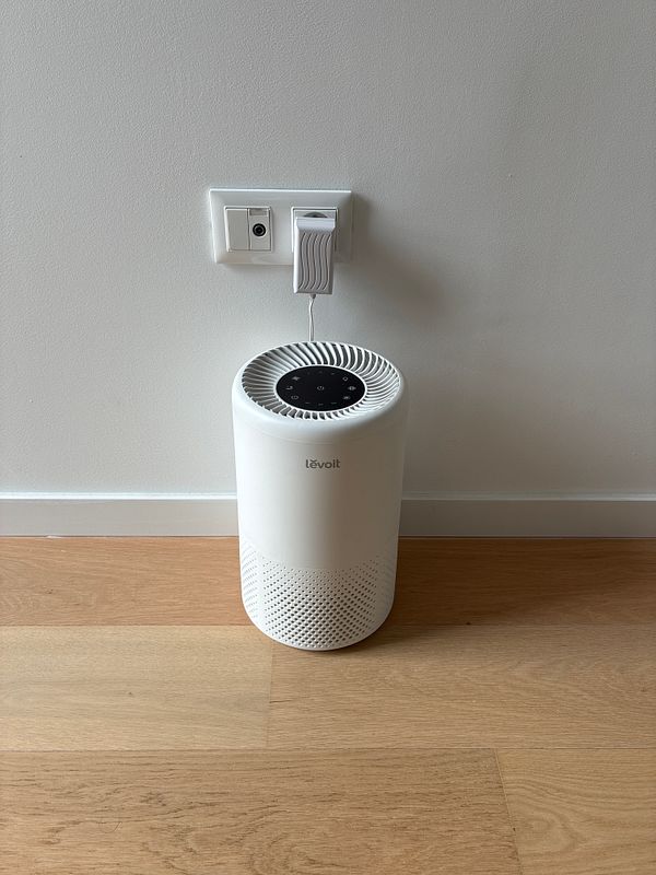 A white Levoit HEPA air purifier plugged into a wall outlet in a room with light wooden floors and a plain white wall.