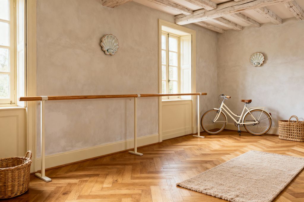 A sunlit French Country home gym with a wall-length ballet barre uses a solid wood rail on cream metal supports against soft greige limewashed plaster walls. Tall cream-trimmed casement windows illuminate herringbone oak parquet flooring beneath whitewashed ceiling beams. Two scallop-shaped wall accents flank the central window. A woven basket rests by the window, another in the corner beside a cream bicycle with brown seat and grips, and a beige textured rug defines a stretching zone.