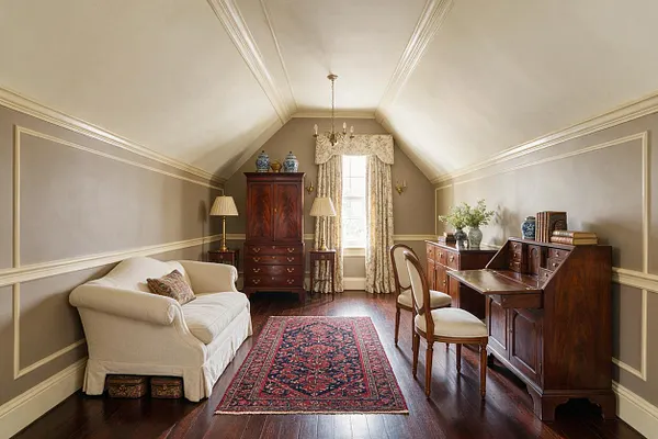 A traditional studio apartment with a sloped ceiling, taupe walls, white crown molding, and dark wood floors. The room contains an upholstered cream sofa, a red patterned area rug, a wooden secretary desk, a tall armoire, two table lamps, a chandelier, and floral drapes framing a central window.