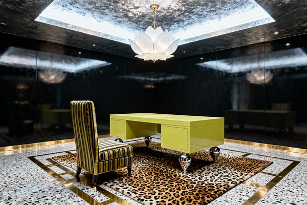 A dramatic Hollywood Regency home office features a chartreuse lacquered desk on polished chrome ball legs centered on a leopard-print rug framed by brass inlays on terrazzo flooring. A high-backed striped velvet side chair sits at the desk, while glossy black lacquer wall panels and gold base trim encircle the room. Overhead, a white lotus-petal chandelier hangs within a silver-leaf coffer with concealed lighting, creating reflective highlights that emphasize the room’s bold contrasts and glamorous materials.