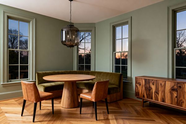 Dining nook with sage green walls and tall double-hung windows surrounding a round oak pedestal table. A curved banquette upholstered in olive velvet wraps the corner, paired with two caramel leather armless chairs on a chevron oak floor. Overhead hangs a smoked-glass globe pendant with a black metal candelabra. A low walnut sideboard with striking grain stands along the right wall. The scene is warmly lit by sunset outside the windows.