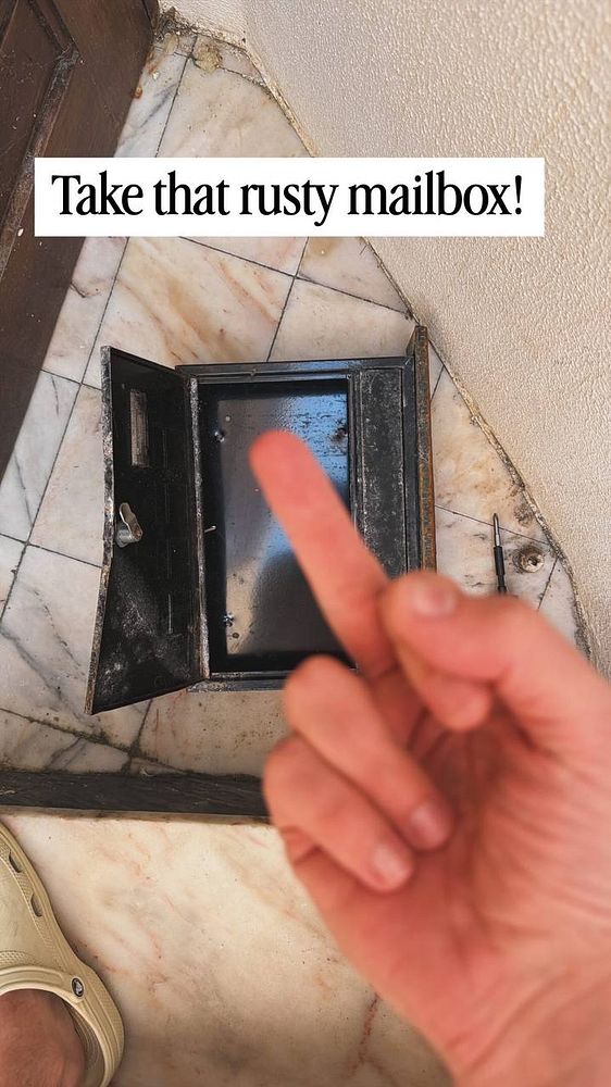An open rusty mailbox is on the ground with a hand showing a middle finger in front of it.