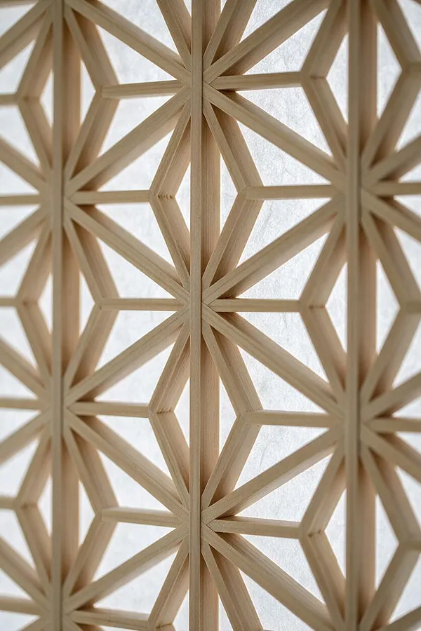 A close-up of a Japanese-style wooden lattice screen with repeating geometric star and hexagon-like patterns. Light natural wood slats are arranged over a softly glowing white translucent background, creating a calm, refined Zen look.