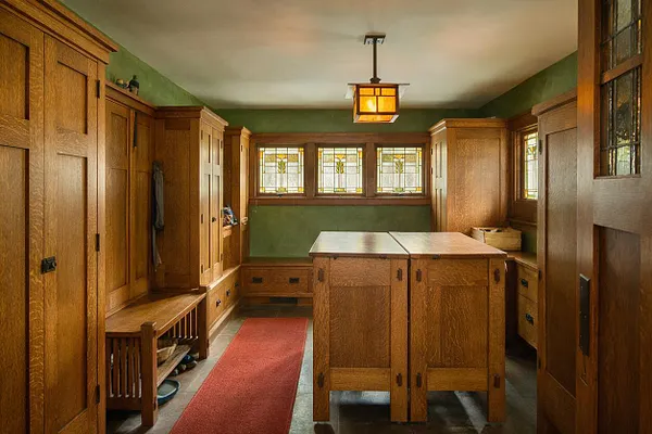 A Craftsman-style laundry room with extensive built-in oak cabinetry, a central work island, and a long red runner rug on a stone floor. Green walls, a warm lantern-style ceiling light, and stained-glass windows create a cozy, historic atmosphere.