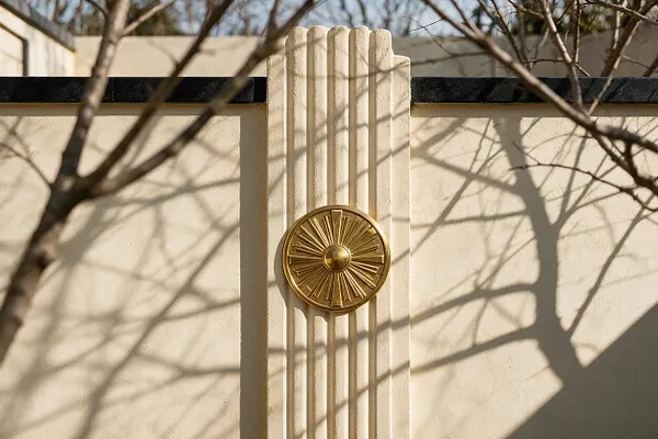 An Art Deco exterior wall in pale cream stucco features a vertical fluted central pilaster and a round brass sunburst medallion. A dark coping caps the wall, while bare tree branches cast soft shadows across the surface in warm daylight.