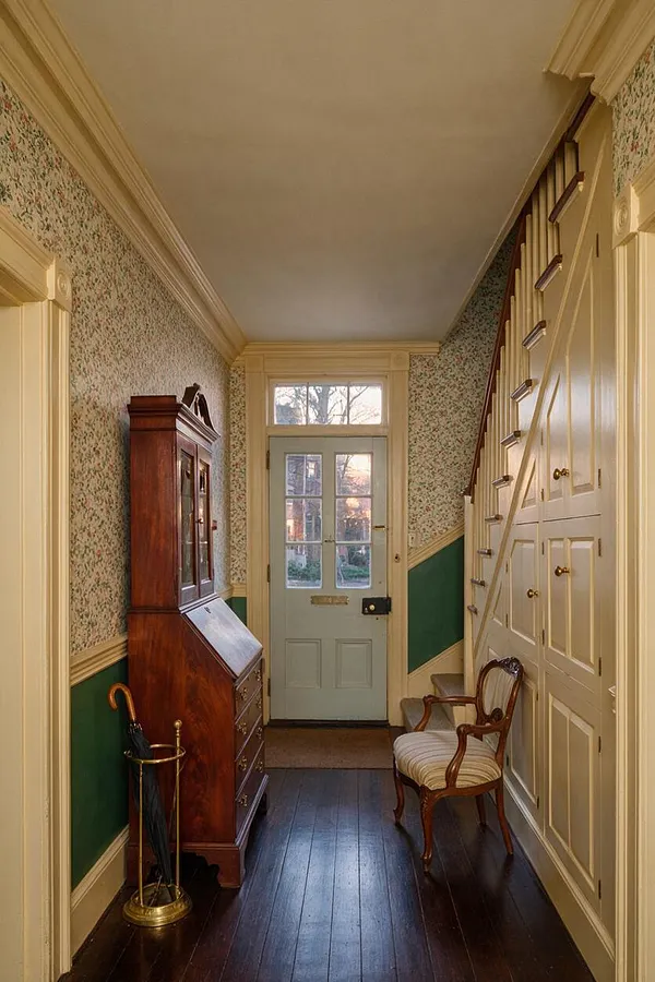 A narrow traditional entryway with floral wallpaper, dark wood floorboards, and cream trim. The space includes a tall mahogany cabinet, a vintage upholstered chair, a brass umbrella stand, and a built-in staircase storage wall beside a pale blue front door.
