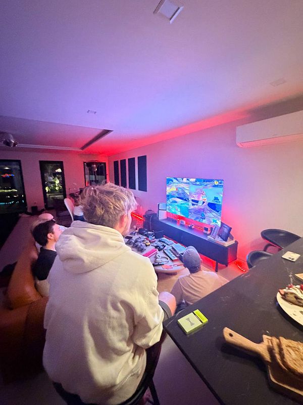 A group of four people playing a multiplayer racing video game in a modern living room with red mood lighting.