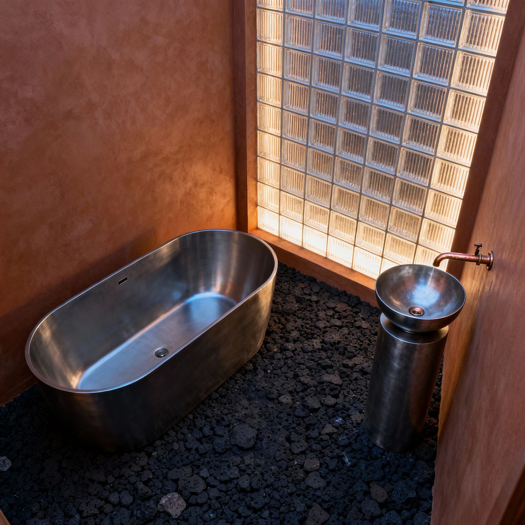 A minimalist bathroom shows a brushed stainless steel freestanding tub and matching pedestal sink placed on a floor of rough black lava rock. Warm terracotta plaster walls surround a full-height glass-block partition that is softly backlit, and a simple wall-mounted copper faucet completes the sparse, spa-like scene with clean lines and sculptural forms.
