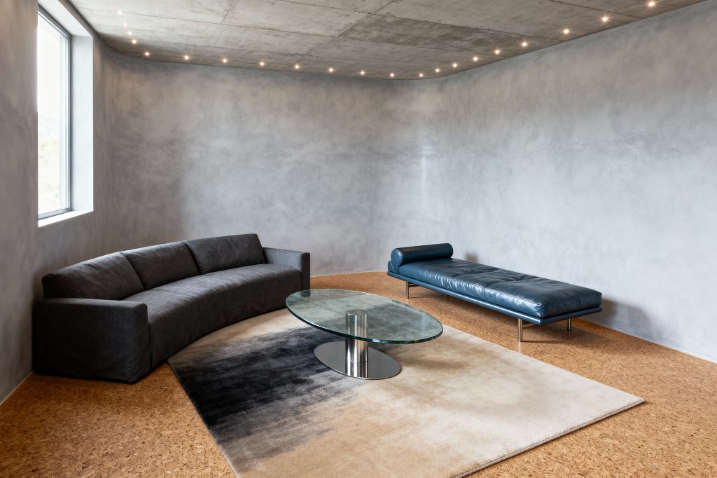 A contemporary living room features a curved charcoal sofa and a streamlined blue leather daybed on slim stainless legs, arranged around an oval glass coffee table with a metal pedestal atop a gradient beige-to-black rug. Warm cork flooring, smooth gray plaster walls, and an exposed concrete ceiling with a ring of small recessed lights frame the space, while a single aluminum-framed window brings in soft natural light.