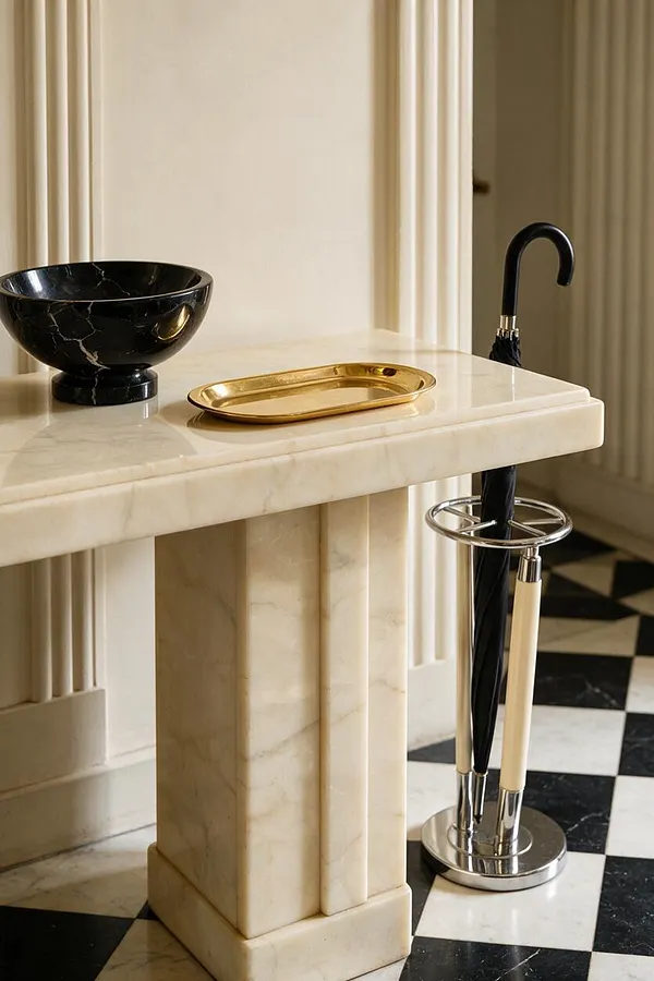 An elegant Art Deco-style entry vignette with a cream marble console table, black marble bowl, and gold tray. A matching umbrella stand holds a black umbrella, and a black-and-white checkered marble floor adds graphic contrast.