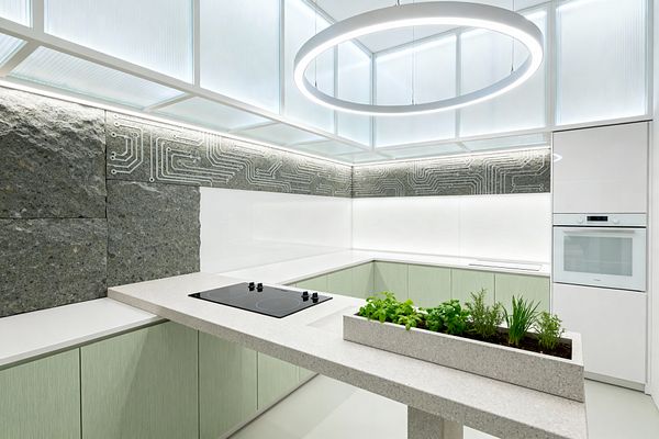A bright futuristic kitchen with a U-shaped layout features pale mint, handleless base cabinets and terrazzo-look countertops with a built-in herb planter and a flush black induction cooktop on the peninsula. The backsplash pairs white back-painted glass with rough gray stone engraved in a circuit-board pattern, while a translucent backlit ceiling and a large circular LED pendant cast cool, even light beside a white built-in wall oven.
