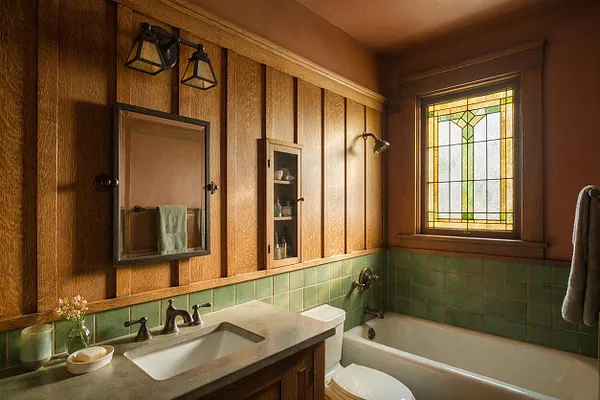 A Craftsman-style bathroom with vertical wood paneling, a green tiled tub surround, and a stained glass window. The space includes a concrete-look vanity top with an undermount sink, dark metal fixtures, a wall mirror, and warm amber lighting.
