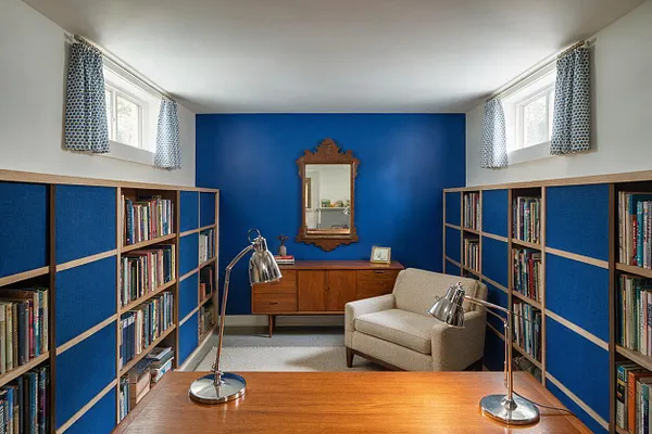 A compact basement library or study with vivid blue built-in bookshelves lining both side walls, filled with books and divided by light wood framing. The back wall is painted deep cobalt blue and features a carved wood mirror above a mid-century style wooden cabinet. A beige armchair sits to the right, while two chrome desk lamps rest on a wooden table in the foreground. Small basement windows near the ceiling are dressed with patterned curtains, and the room has a bright, cozy atmosphere.