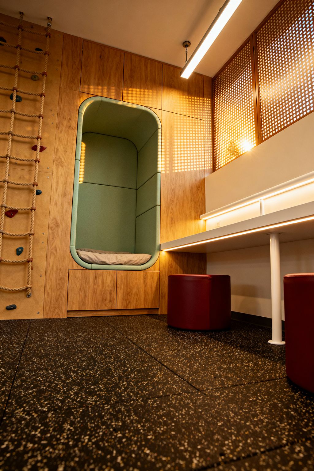 Modern children’s room with a built-in oak-veneer wall featuring a rounded, mint-green cushioned reading alcove above low drawers. To the left, a rope ladder and small climbing holds are mounted on the wooden panel. On the right, a long white desk with under-shelf LED lighting stands on a white metal leg, paired with two burgundy cylindrical stools. Perforated wood screens filter warm sunlight, and a black speckled rubber floor provides durable, safe footing.