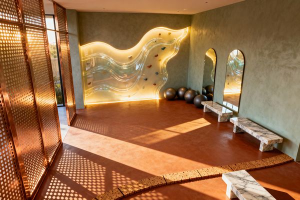 A sunlit home gym with a translucent, wavy acrylic climbing wall backlit by warm LEDs, set against sage green plaster walls. Perforated copper screens flank a window, casting dotted shadows across a terracotta microcement floor. Three arched mirrors with brass trim and white marble benches line the right wall, charcoal exercise balls sit below, and a curved band of cork pavers arcs across the open space.