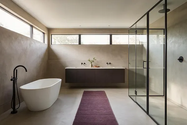 A modern master bathroom with a freestanding white bathtub on the left, a black floor-mounted tub filler, a dark floating vanity along the back wall, and a glass-enclosed shower on the right. Beige plaster walls, a light concrete floor, narrow clerestory windows, and a purple runner add warmth to the minimalist space.