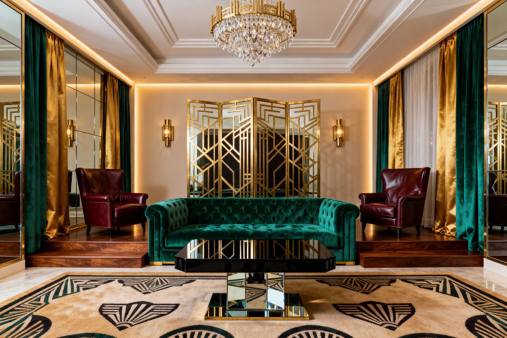 Symmetrical media room with an emerald tufted velvet sofa centered between two burgundy leather armchairs set on raised walnut platforms. Behind, a mirrored folding screen with gilded Art Deco geometry and brass sconces glows under perimeter LED lighting, while a crystal chandelier crowns a coffered ceiling. A black lacquer coffee table with mirrored base sits on a cream rug with emerald and black deco motifs, framed by teal and gold velvet drapery and mirrored wall panels.