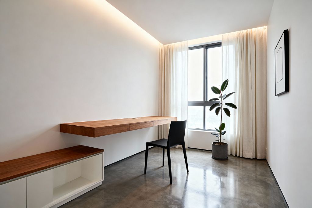 A minimalist home office features a wall-mounted walnut desk with slim drawers and a single black chair on a polished concrete floor. A large gray metal-framed window with floor-length sheer curtains brings diffused daylight, while warm cove lighting grazes smooth white walls; a low white credenza with a walnut top, a potted rubber plant, and a simple black-framed line print complete the uncluttered space.