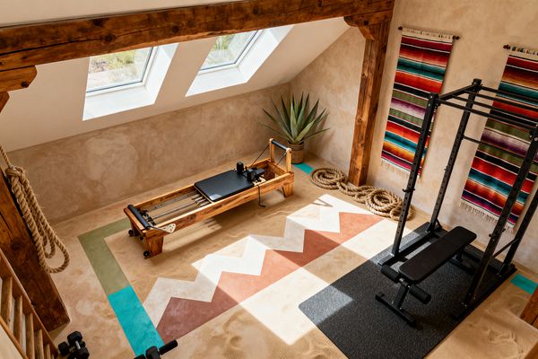 A sunlit attic home gym features two roof skylights, exposed heavy wood beams, and sand-colored plaster walls. A wood Pilates reformer sits beside a black steel squat rack on a speckled rubber mat, with a painted zigzag floor pattern in terracotta, cream, and turquoise, colorful serape wall hangings, an agave plant in a woven basket, coiled battle ropes and a hanging climbing rope, plus small black dumbbells arranged near the railing.