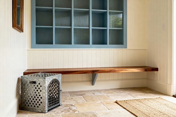 A farmhouse mudroom with cream vertical beadboard features a floating walnut bench supported by a blue bracket beneath built-in dusty-blue cubbies with reeded glass. The floor is warm beige stone tile with a small woven jute rug. A galvanized metal dog crate sits at the bench’s end and a wood-framed mirror hangs on the wall. Soft daylight from the right brightens the space and highlights the natural textures and calm palette.