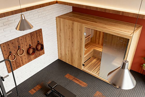 A Mid-Century styled home gym features a built-in oak sauna with slatted benches, a clear glass door, and a frosted side panel. A white-painted brick wall holds a walnut pegboard with wooden rings on leather straps; black coin-pattern rubber flooring with wood inlays anchors a black weight bench, while a terracotta accent wall, warm cove lighting, brushed-metal cone pendants, and a small plant complete the clean, inviting look.