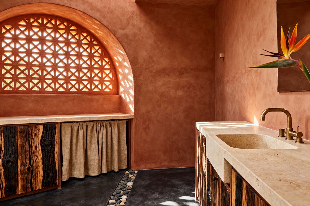 A biophilic laundry room features terracotta plaster walls and a large arched lattice window that casts patterned sunlight across the space. A pale stone countertop with an integrated sink and brass faucet pairs with rustic cabinetry in charred wood and a natural linen skirt; dark concrete floors include a pebble-lined channel, while a bird-of-paradise stem brings a vivid botanical note.