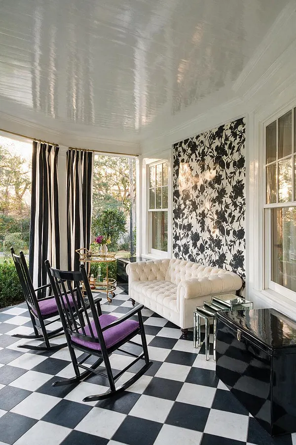 A stylish front porch with a glossy white ceiling, black-and-white checkerboard floor, and large windows framing garden views. Black rocking chairs with purple cushions sit near a tufted white sofa, mirrored side tables, and bold black-and-white floral wall covering, creating a glamorous Hollywood Regency look.