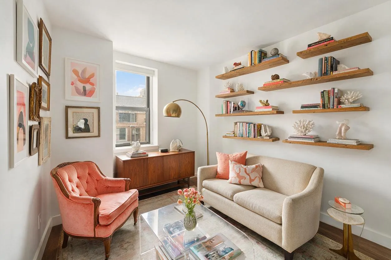 A bright eclectic media room with a coral velvet armchair, a cream sofa, floating wooden shelves filled with books and sculptural decor, and a walnut credenza beneath a window. Framed wall art, a brass arc floor lamp, a clear acrylic coffee table, and layered neutral and blush tones create a curated, airy feel.