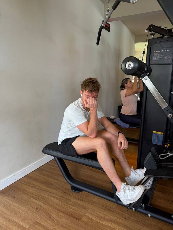 A photo of a woman in a gym lifting a barbell in an overhead press position, wearing a red sports bra and black shorts, in a gym setting with some workout equipment in the background.