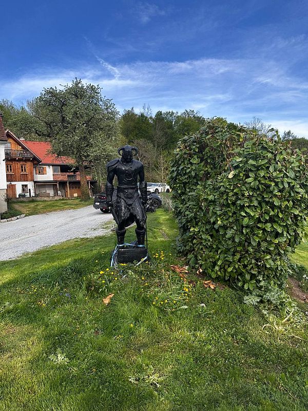 A black statue of a warrior stands in a grassy area near a house and a hedge.