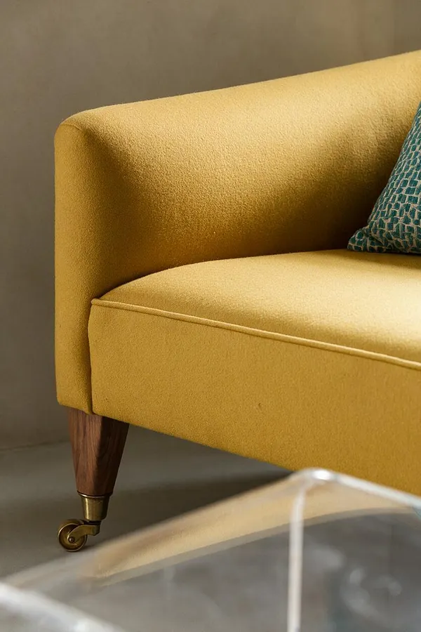 A close-up of a mustard yellow upholstered sofa shows a smooth curved arm, cushioned seat, and a tapered walnut leg with a brass caster. A teal patterned pillow peeks in at the edge, while a transparent glass table blurs in the foreground against a warm neutral wall.