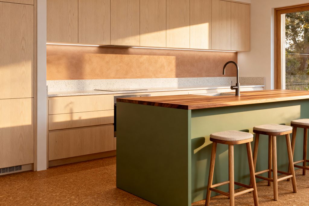 A sunlit Scandinavian kitchen shows flat-front light oak cabinets, a speckled light terrazzo countertop, and a terracotta-toned plaster backsplash illuminated by a slim under-cabinet light. A sage green island with a thick butcher-block wood top holds a brushed stainless faucet and undermount sink, with three wooden stools upholstered in beige fabric. Natural cork flooring and a wood-framed window complete the calm, minimal space featuring clean lines and warm materials.