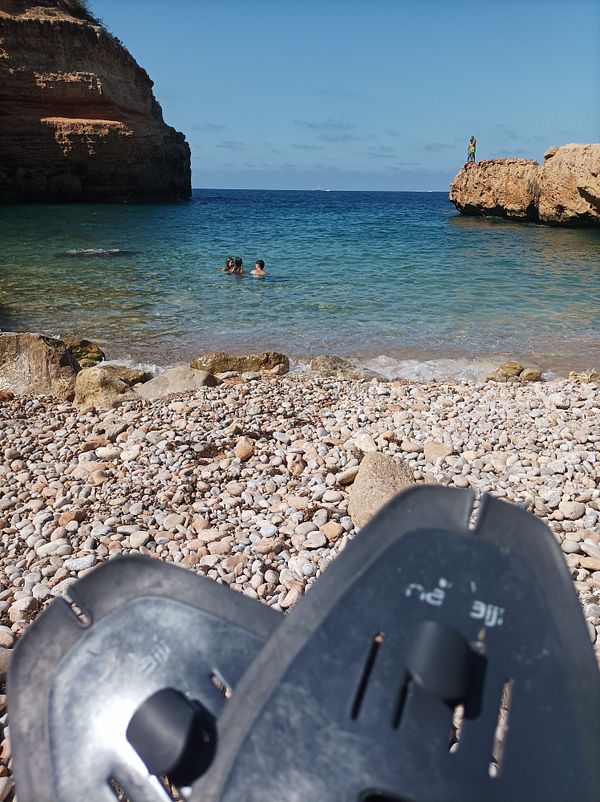 A scenic beach view with swimmers and a person on a rock, featuring swimming fins in the foreground.