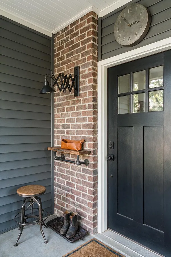 A front porch styled in industrial decor features a black paneled door with small window panes, a red brick wall, and dark gray horizontal siding. A black metal wall sconce, round wall clock, rustic stool, small shelf with a leather pouch, and a boot tray with boots complete the entryway.