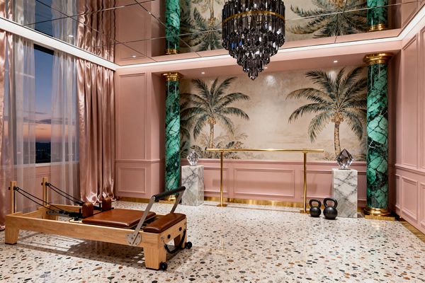 Luxurious home gym with blush pink paneled walls and a large palm-tree mural, flanked by emerald green marble columns with gold capitals and a brass ballet barre beneath a mirrored ceiling and smoky crystal chandelier, all set over terrazzo floors. At left, floor-to-ceiling satin drapes and sheers frame a sunset view near a wood Pilates reformer with brown leather, while white marble pedestals with crystal sculptures and two black kettlebells accent the right.