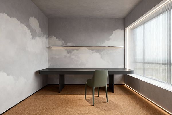 A minimalist home office with a wall-to-wall graphite desk with rounded corners and central supports, paired with a muted olive chair on cork flooring. Soft gray walls display a cloud mural; above the desk a slim floating metal shelf with integrated LED casts an even glow. A full-height window with fluted glass and perimeter floor lighting brightens the serene space, complemented by small recessed ceiling downlights.