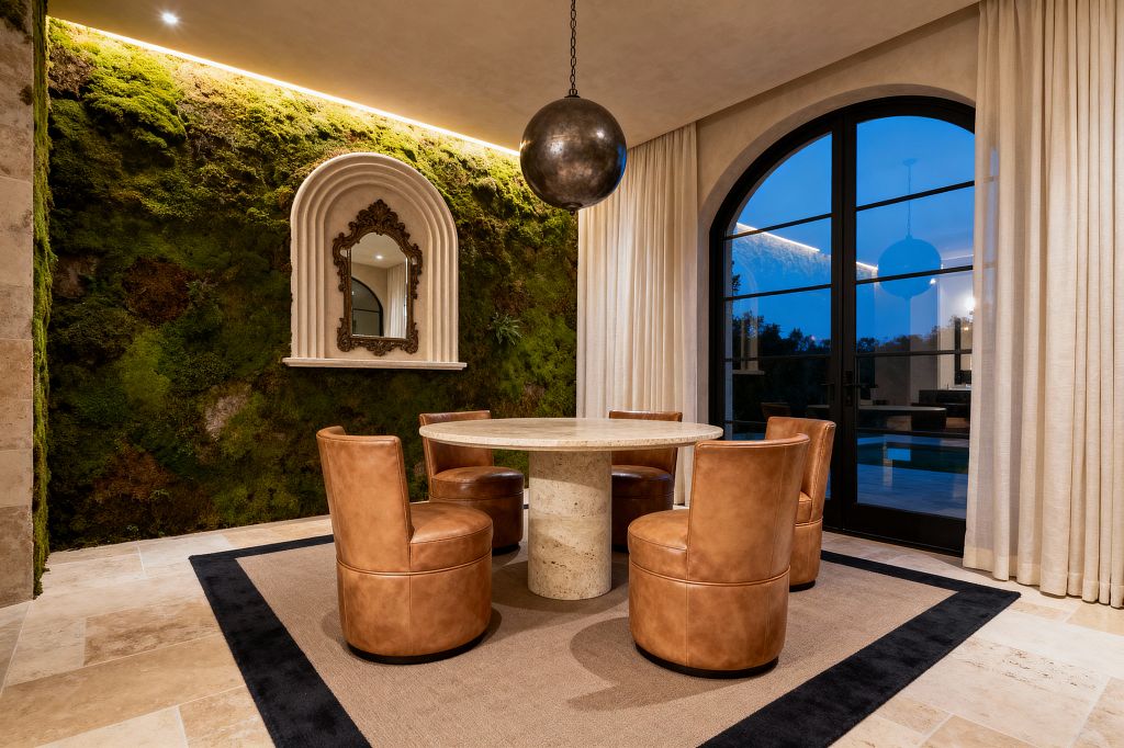 A transitional dining room featuring a round travertine pedestal table surrounded by four cognac leather barrel chairs on a light rug with a dark border. A full moss-covered accent wall with warm cove lighting includes an arched plaster niche holding an ornate mirror. To the right, a large arched black metal-framed glass door is flanked by floor-length ivory linen curtains, while a dark spherical pendant hangs above and beige stone tiles cover the floor.