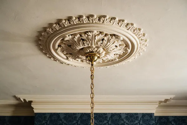 An ornate Victorian-style ceiling detail with a carved plaster medallion featuring layered oval moldings and leaf motifs. A brass ceiling rose at the center supports a hanging chain, with cream crown molding and deep blue patterned wallpaper below.