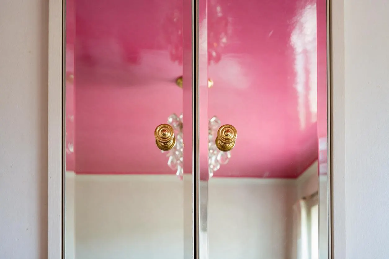 A glossy pink cabinet with mirrored doors fills the frame, each door fitted with a round brass knob. The reflective surface shows a bright room with white walls, a pink ceiling, and a crystal chandelier, creating a glamorous Hollywood Regency look.