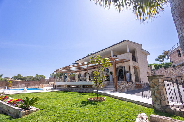 A luxurious Mediterranean-style villa with white walls, blue shutters, and a swimming pool surrounded by palm trees and a scenic mountain backdrop.