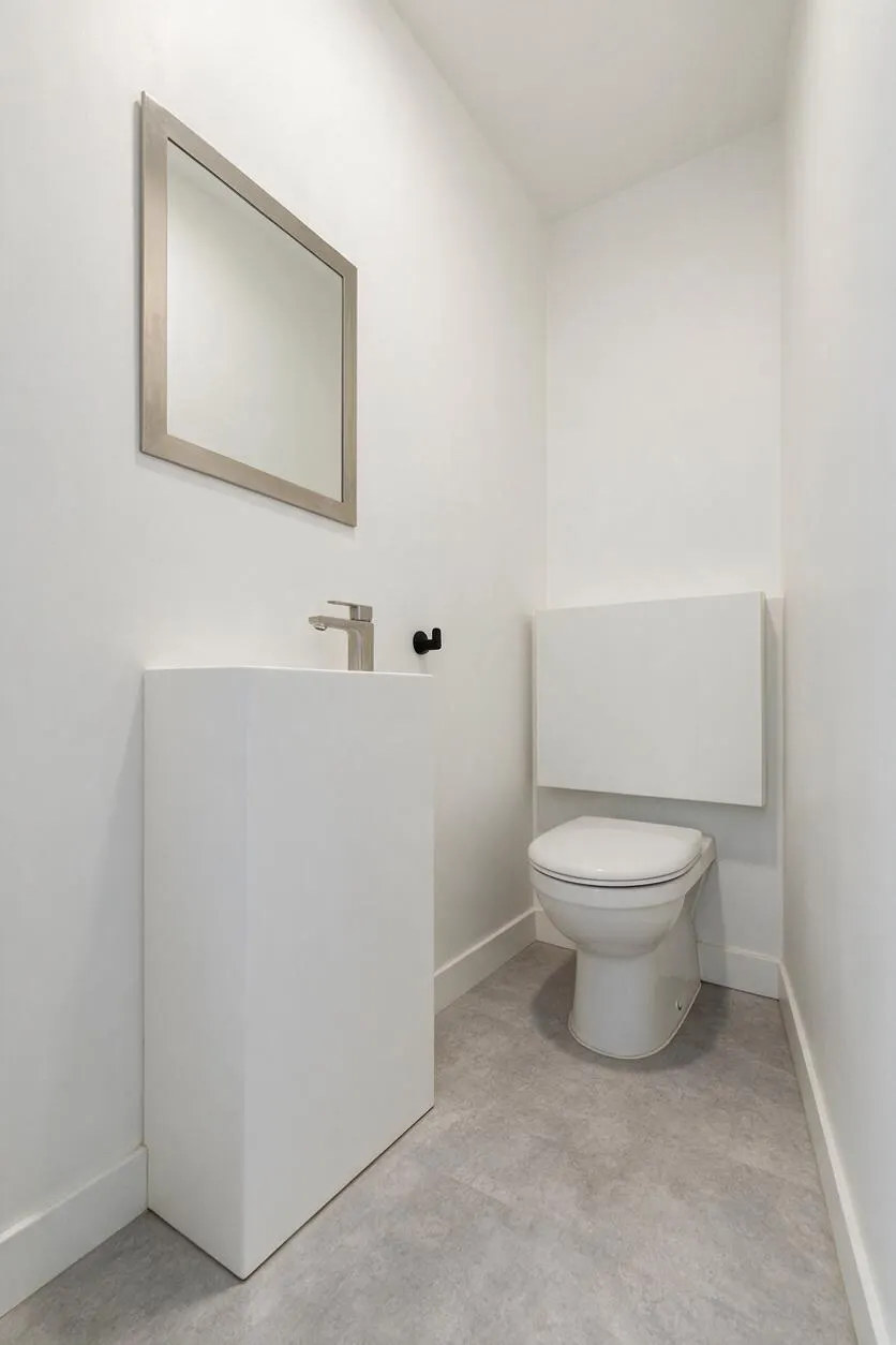 A compact powder room with white walls, a slim framed mirror, a minimalist white vanity with a simple chrome faucet, and a wall-mounted toilet. The space has light gray flooring and a clean, uncluttered modern look.