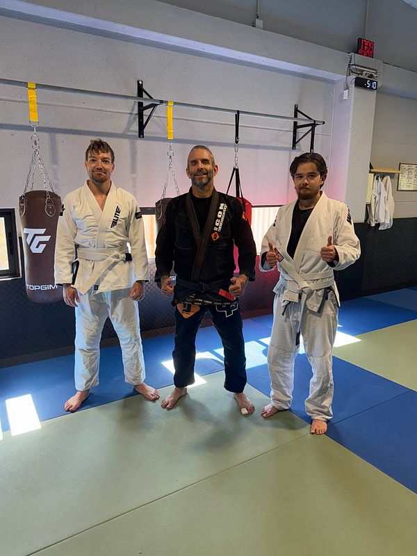 Three individuals pose together in a Brazilian Jiu-Jitsu training environment.