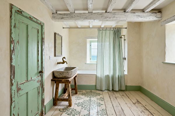 An airy shabby chic bathroom features a distressed mint-green door, matching skirting and window trim, and whitewashed ceiling beams over warm cream plaster walls. A carved stone trough sink sits on a rustic trestle table beneath a weathered wood mirror, with a brass swan-neck wall spout. Lime-washed plank floors carry sage stenciling, while a green-and-white striped cotton curtain softens a tub with exposed brass shower beside a small sea-facing window.