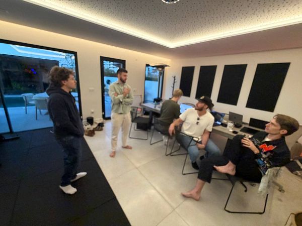 Five young men are casually interacting in a modern coworking space featuring desks with laptops and minimalistic decor.