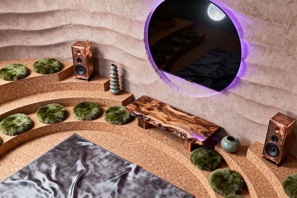 A surreal media room with stepped cork amphitheater seating arranged before a circular screen set into a rippled plaster wall, glowing with purple light and reflecting a moon. Moss-green faux fur cushions line the steps, flanked by hammered copper speakers; a live-edge walnut bench, a stack of turquoise glazed ceramic vases, and a small round pot rest on the tiers, with a silver satin throw spread across the floor.