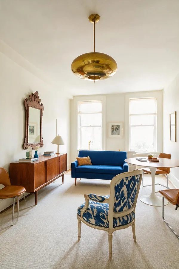 A bright eclectic home office with white walls, a large brass pendant light, and a mix of vintage and modern furnishings. The room includes a blue sofa, a carved wood mirror above a walnut cabinet, a round pedestal table, and upholstered accent chairs in tan and blue patterned fabric.