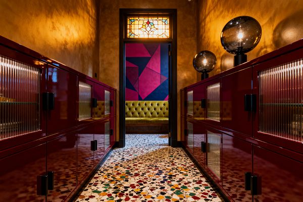 A narrow, symmetrical mudroom features high-gloss burgundy cabinets with fluted-glass doors on both sides and black pulls, set on a white terrazzo floor speckled with multicolored chips. Warm golden plaster walls and two smoked-glass globe lamps create moody light. A doorway with a stained-glass transom reveals an olive green tufted bench and a bold magenta and navy geometric wall beyond.