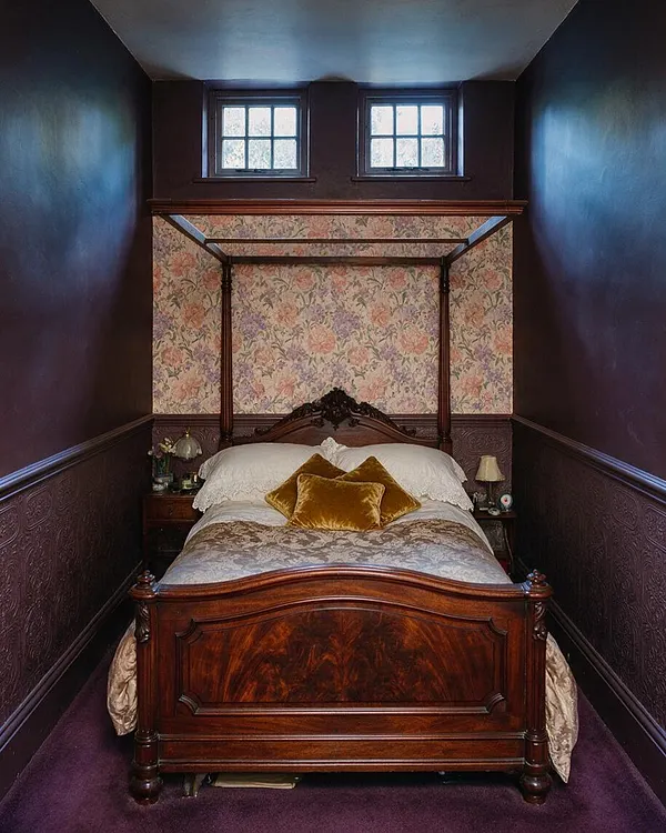 A narrow Victorian-style basement bedroom with dark plum walls, ornate textured wainscoting, and a rich wood canopy bed centered beneath two small windows. Floral wallpaper fills the headboard wall, while white bedding and gold velvet pillows soften the deep, moody palette.