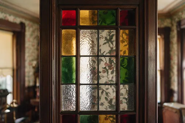 A close-up of a Victorian-style wooden door or partition with a leaded stained glass insert. The glass features textured clear panes framed by deep brown wood and accented with jewel-tone red, green, and amber sections, with a softly patterned vintage room blurred in the background.