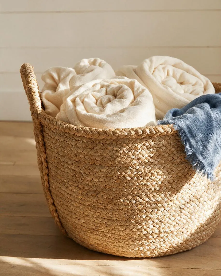 A round woven seagrass basket sits on a light wood floor, filled with rolled cream-colored blankets and draped with a faded blue throw. The neutral palette and natural textures create a relaxed coastal feel.
